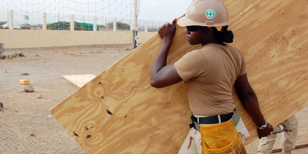 A construction worker wearing a tan shirt, camouflage pants, a hard hat, and sunglasses carries a large sheet of plywood at a work site. The worker has a yellow tool belt and is walking across a sandy area with a net and fencing in the background.